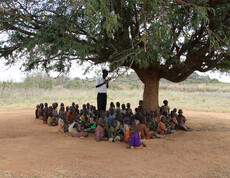 Im Schatten eines Baumes in Uganda werden viele Kinder von einem Lehrer unterrichtet.