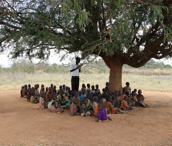 Im Schatten eines Baumes in Uganda werden viele Kinder von einem Lehrer unterrichtet.