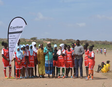 Eine Gruppe junger Frauen in roten Sporttrikots steht gemeinsam mit Erwachsenen und einem Banner am Spielfeldrand. Sie nehmen an einem Friedensprojekt teil, das Sport und Aufklärungsarbeit verbindet.