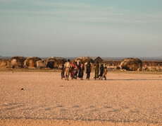 Eine Gruppe von Menschen geht über eine trockene, karge Ebene in Richtung traditioneller Rundhütten. Die Szene zeigt ein Nomadenlager in einer von Dürre betroffenen Region.