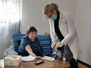 Eine Krankenschwester mit Maske misst Blutdruck bei einer Dame, die auf einer blauen Couch sitzt.