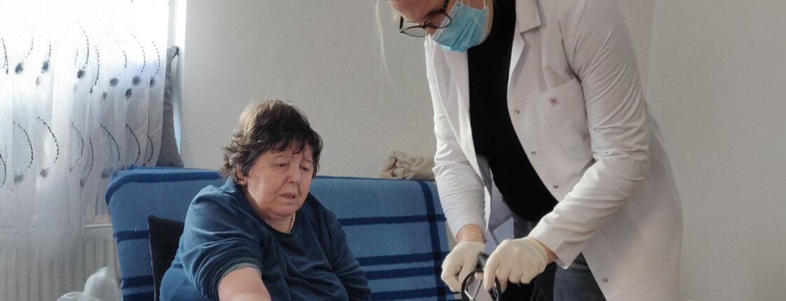 Eine Krankenschwester mit Maske misst Blutdruck bei einer Dame, die auf einer blauen Couch sitzt.