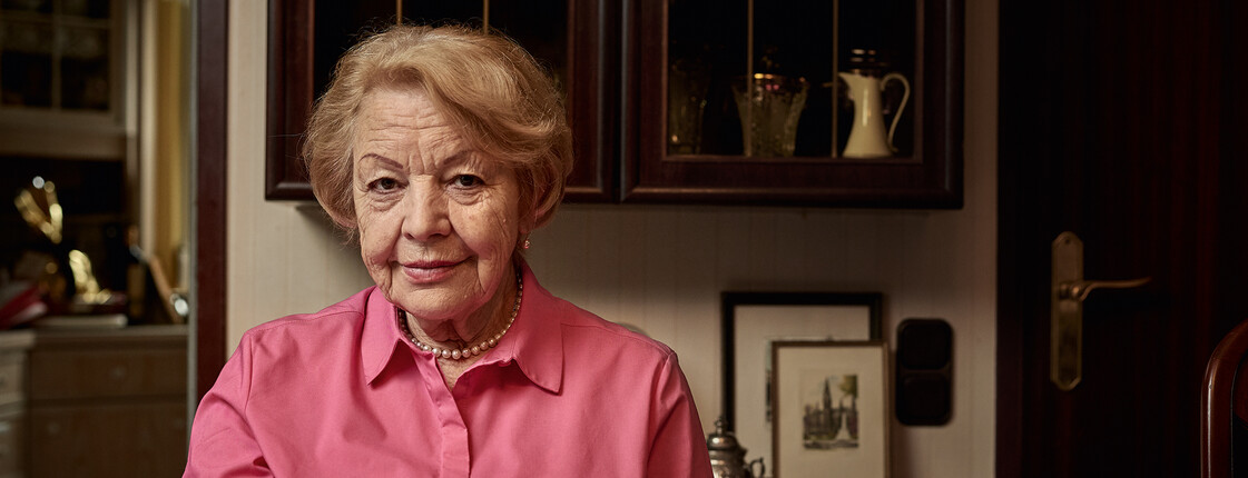 Eine ältere Dame sitzt bei einer Tasse Tee am dunklen Esstisch und lächelt.