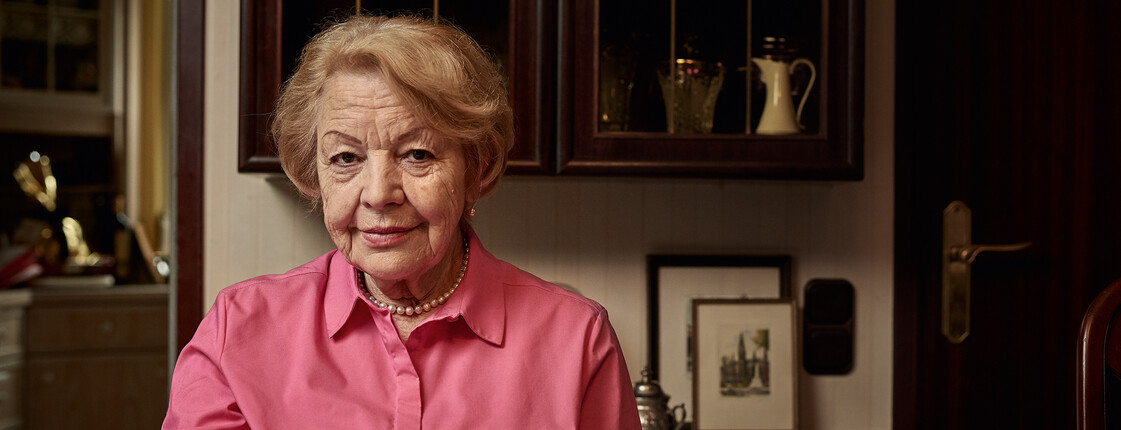 Eine ältere Dame sitzt bei einer Tasse Tee am dunklen Esstisch und lächelt.