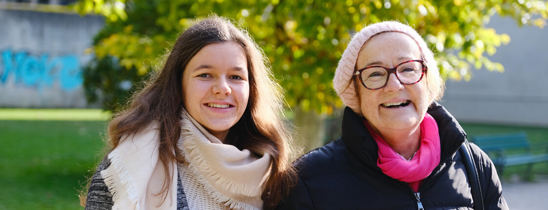 Eine junge Frau begleitet eine Seniorin, beide tragen Winterkleidung, beim Spazierengehen durch einen Park.