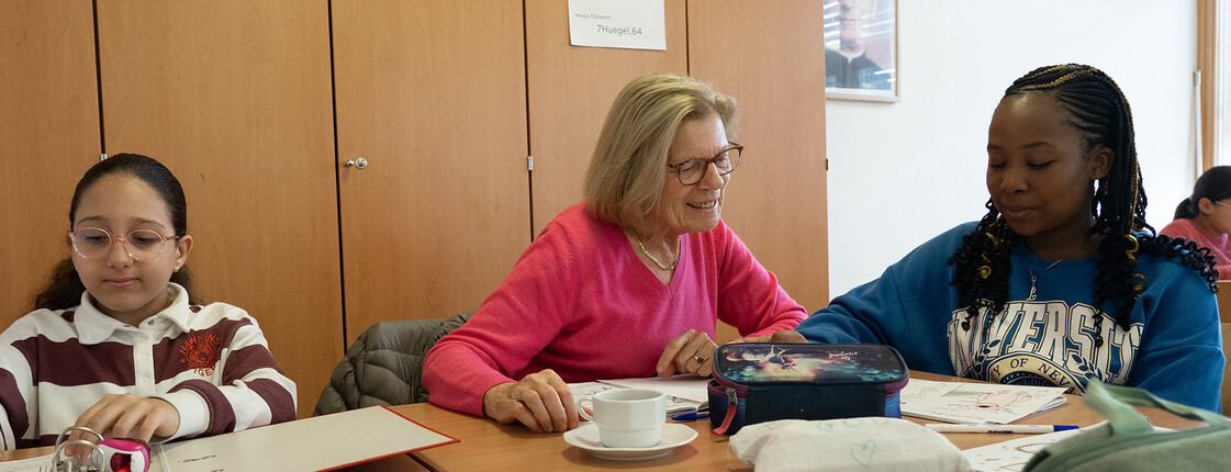 Kinder arbeiten an Heften und Arbeitsblättern, eine Freiwillige begleitet das Lernen.
