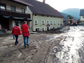Zwei Caritas-Mitarbeiter gehen über eine verschlammte Straße nach einem Hochwasser.
