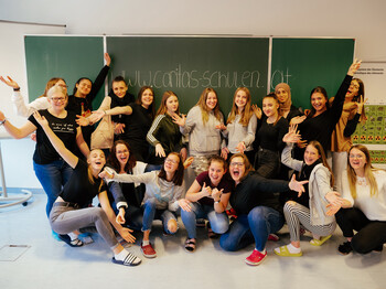Auf der Tafel, vor der sich viele Schüler*innen der Caritas HLW in Klagenfurt versammelt haben, sich freuen und die Hände in die Luft strecken, steht die Website www.caritas-schulen.at.