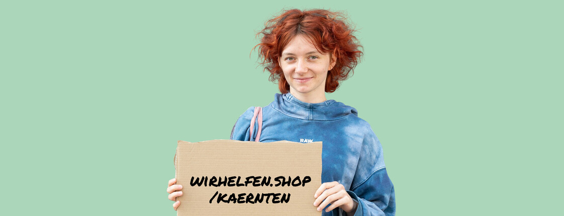 Eine junge, rothaarige Frau hält ein Schild in der Hand auf dem wirhelfen.shop/kaernten geschrieben steht.