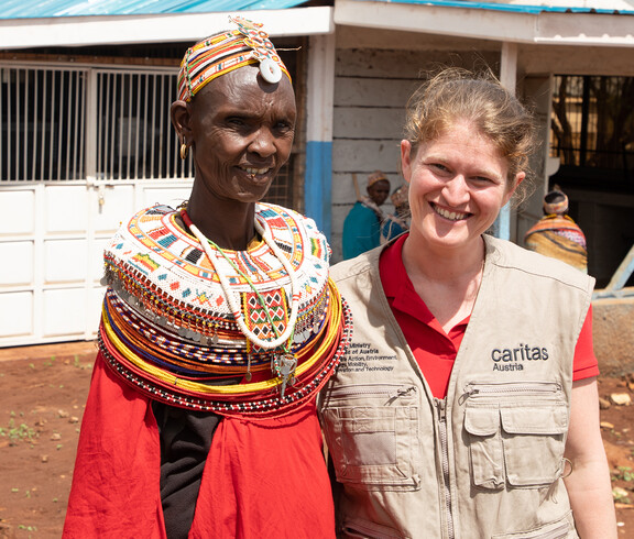 Caritas-Projektreferentin Theresa Sacher steht gemeinsam mit einer afrikanischen Frau in Marsabit. Beide lächeln, im Hintergrund sind weitere Personen und Gebäude zu sehen. Die Szene zeigt den direkten Austausch bei einem Projektbesuch.