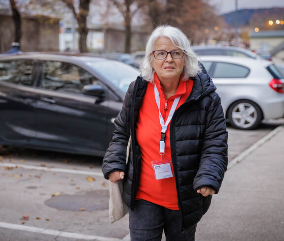 Margit Müller geht durch einen Parkplatz mit Tasche und rotem Shirt unter schwarzer Jacke