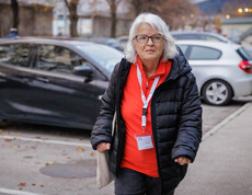 Margit Müller geht durch einen Parkplatz mit Tasche und rotem Shirt unter schwarzer Jacke