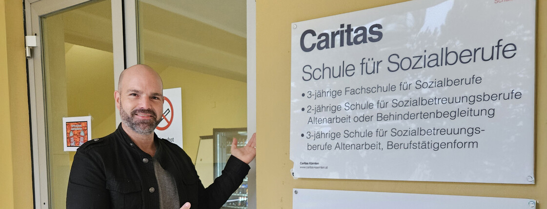 Schulleiter steht vor Eingang der Caritas-Schule für Sozialberufe in Wolfsberg und zeigt auf das Schulschild.
