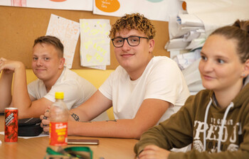Zwei Schüler sitzen im Klassenzimmer und lächeln in die Kamera. Eine Schülerin sitzt noch daneben und sieht zur Tafel.