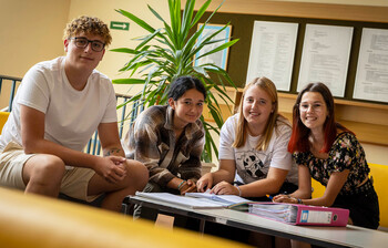 Vier Schüler*innen sitzen auf einem gelben Sofa, haben Schulmaterial vor ihnen liegen und lächeln in die Kamera.