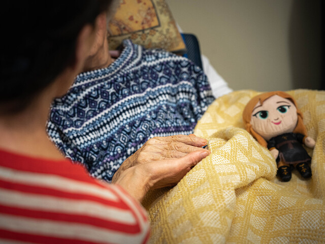 Eine ältere Person im Pflegebett wird von einer Angehörigen gehalten, eine Stoffpuppe liegt auf der gelben Decke.