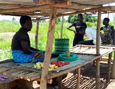 Eine junge Frau sitzt unter einem schützenden Dach bei ihrem eigenen kleinen Markt. Im Hintergrund sind noch weitere Frauen, die ebenfalls einen kleinen Stand betreiben.