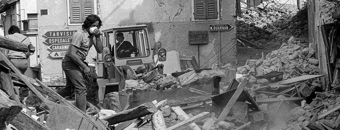 Trümmer bedecken eine Straße, ein Bagger räumt Schutt vor beschädigten Häusern nach dem Erdbeben in Friaul.