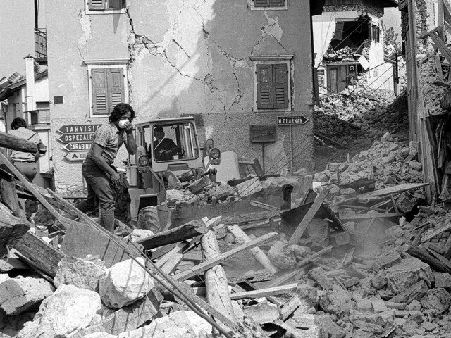 Trümmer bedecken eine Straße, ein Bagger räumt Schutt vor beschädigten Häusern nach dem Erdbeben in Friaul.