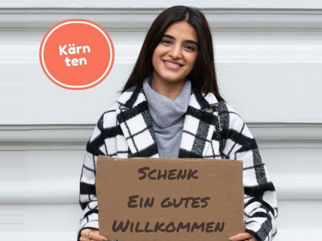 Eine junge Frau lächelnd in die Kamera. Sie hält ein Schild mit "Schenk ein gutes Willkommen".