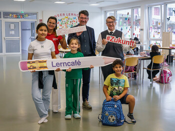 Zwei Männer, eine Caritas-Mitarbeiterin und drei Kinder halten Lerncafé-Schilder in einem Lernraum.