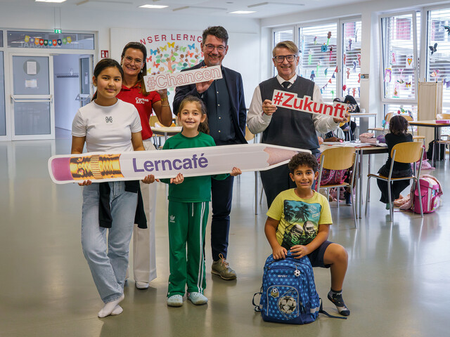 Zwei Männer, eine Caritas-Mitarbeiterin und drei Kinder halten Lerncafé-Schilder in einem Lernraum.
