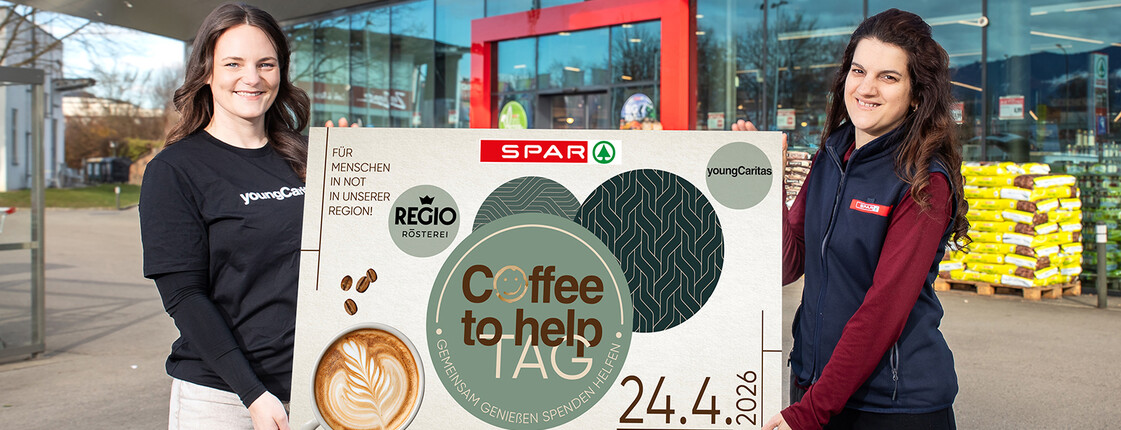 Zwei Frauen halten vor einem SPAR-Markt ein Plakat für den Coffee to help Tag am 24.4.2026 mit youngCaritas.
