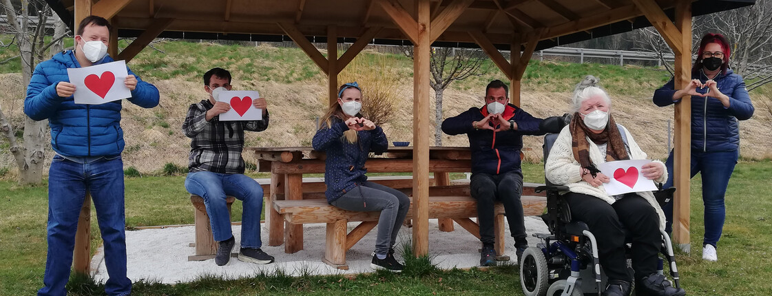 Bewohner*innen unserer Wohnhäuser für Menschen mit Behinderung unserers Caritas: Team Lebensgestaltung stehen mit Betreuer*innen am und im Pavillion und halten Schilder mit Herzen entgegeben. Alle tragen Masken, da das Foto zu Corona-Zeiten aufgenommen wurde.