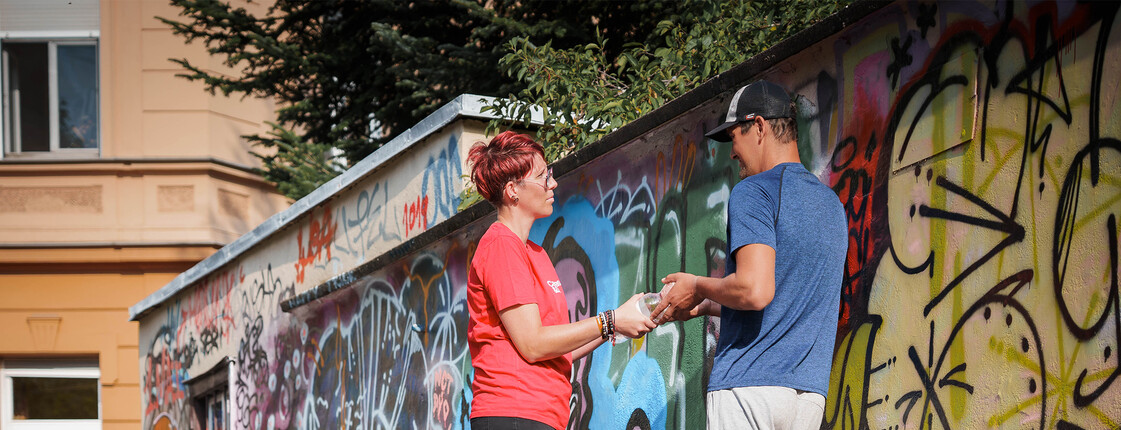 Caritas‑Mitarbeiter*in übergibt einem obdachlosen Menschen Hilfsgüter an einer Graffitiwand im öffentlichen Raum.