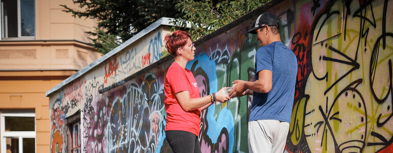 Caritas‑Mitarbeiter*in übergibt einem obdachlosen Menschen Hilfsgüter an einer Graffitiwand im öffentlichen Raum.