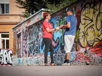 Caritas‑Mitarbeiter*in übergibt einem obdachlosen Menschen Hilfsgüter an einer Graffitiwand im öffentlichen Raum.