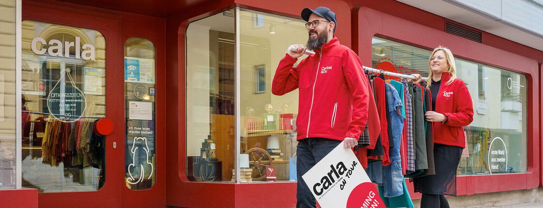 Patrick Pajer, Leiter der carlas, und Christina Staubmann, Bereichsleiterin Beschäftigung und Betriebe, gehen mit einem Schild mit "carla on tour - coming soon" und einer Kleiderstange den Bürgersteig vor dem carla Klagenfurt entlang.