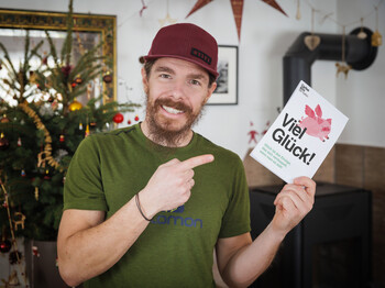 Ein Mann lächelt in die Kamera und hält eine Karte auf der "Viel Glück" steht.