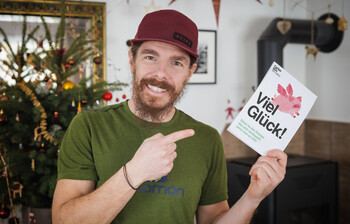 Ein Mann lächelt in die Kamera und hält eine Karte auf der "Viel Glück" steht.