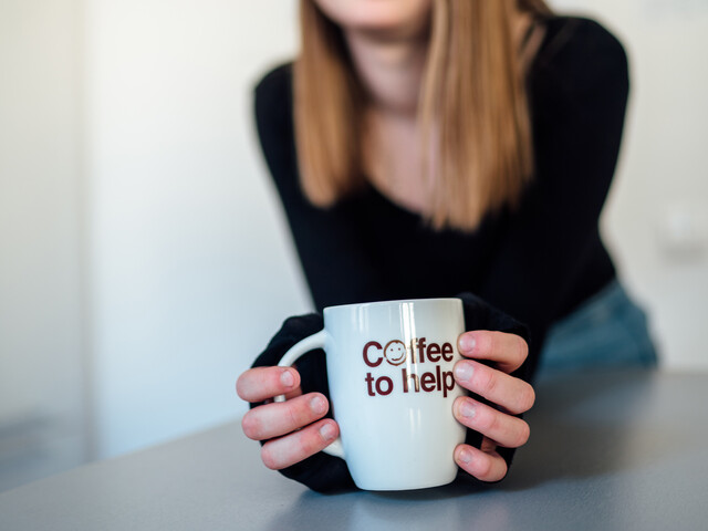 Ein junges Mädchen mit langen hellbraunen Haaren hält eine Coffee to help-Tasse in der Hand.