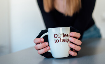 Ein junges Mädchen mit langen hellbraunen Haaren hält eine Coffee to help-Tasse in der Hand.