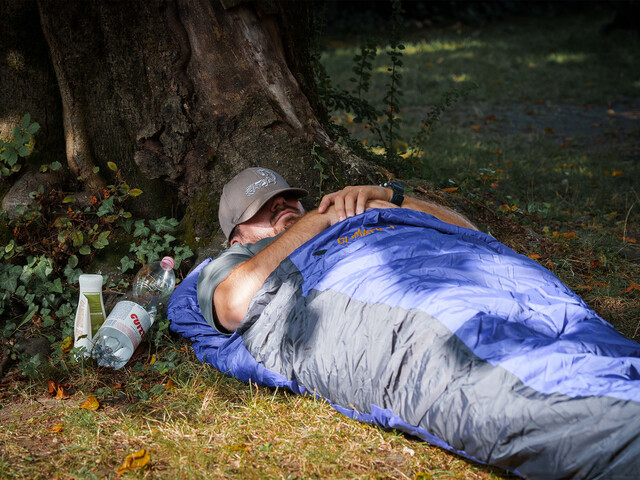 Obdachloser Mensch liegt tagsüber in einem Schlafsack unter einem Baum, Trinkflaschen liegen daneben.