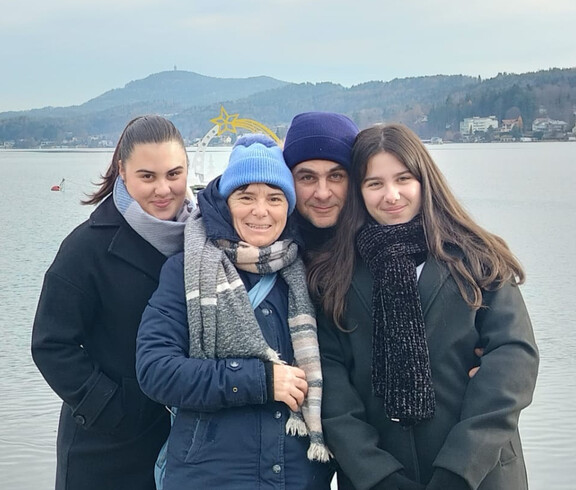 Vier Personen stehen am Ufer eines Gewässers, im Hintergrund Berge und Ortschaft.