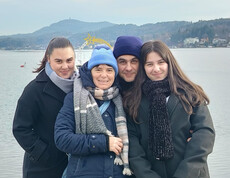 Vier Personen stehen am Ufer eines Gewässers, im Hintergrund Berge und Ortschaft.