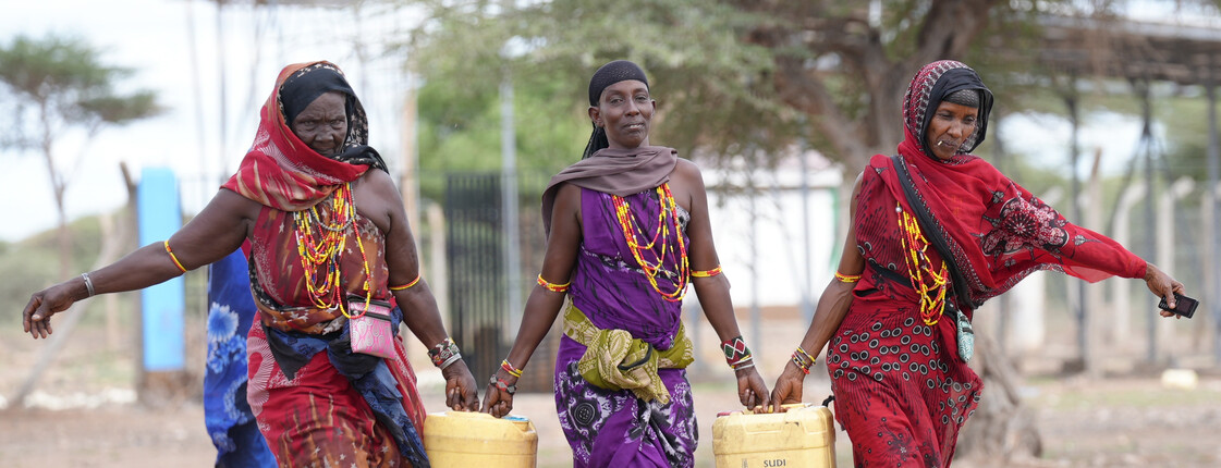 Drei Frauen mit traditioneller Kleidung und Schmuck tragen Wasserkanister in Kenia