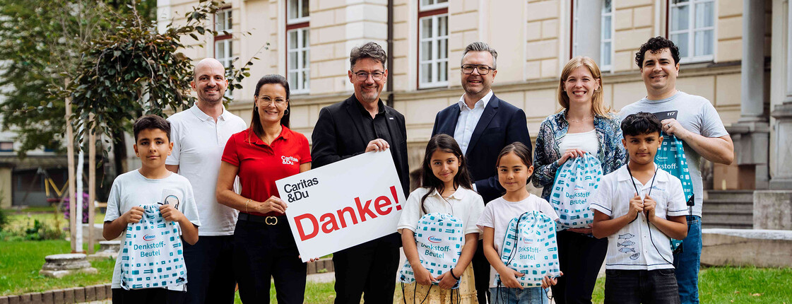 Gruppe mit Kindern und Erwachsenen überreicht Denkstoff-Schulbeutel vor Gebäude; Schild mit Aufschrift „Danke!“.