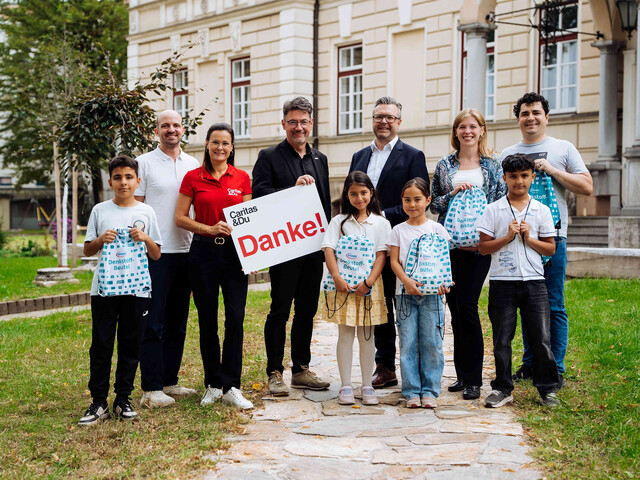 Gruppe mit Kindern und Erwachsenen überreicht Denkstoff-Schulbeutel vor Gebäude; Schild mit Aufschrift „Danke!“.