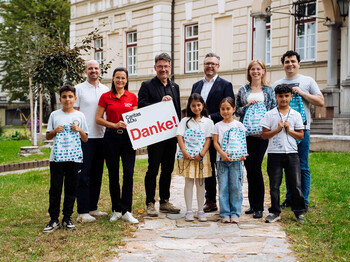 Gruppe mit Kindern und Erwachsenen überreicht Denkstoff-Schulbeutel vor Gebäude; Schild mit Aufschrift „Danke!“.
