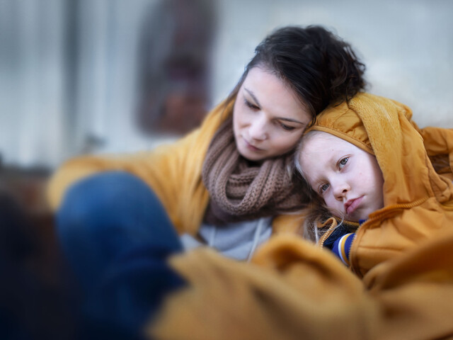 Eine Mutter sitzt mit verzweifeltem Gesichtsausdruck mit ihrem in eine Decke eingewickelten Kind auf einer Couch.