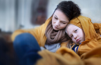 Eine Mutter sitzt mit verzweifeltem Gesichtsausdruck mit ihrem in eine Decke eingewickelten Kind auf einer Couch.