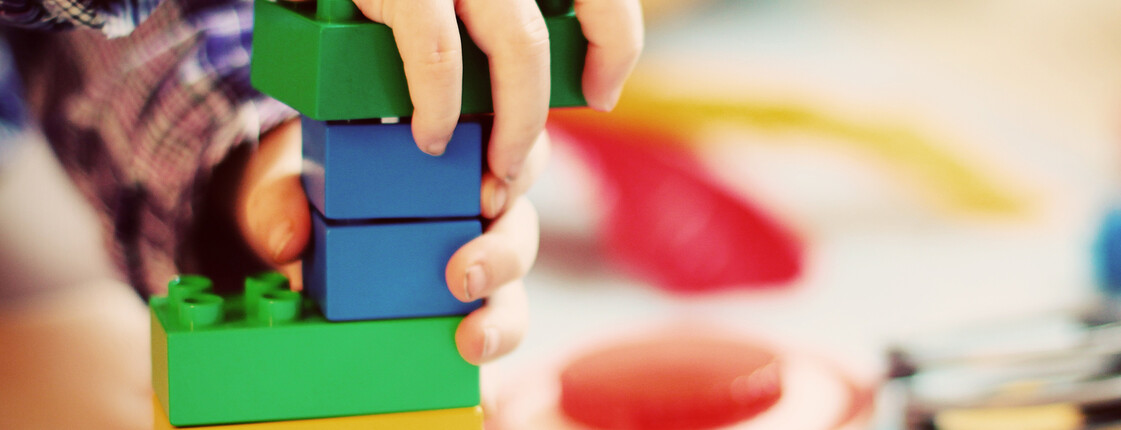 Eine Kinderhand baut mit Bauklötzen einen bunten Turm. Im Hintergrund sieht man weiteres Spielzeug.