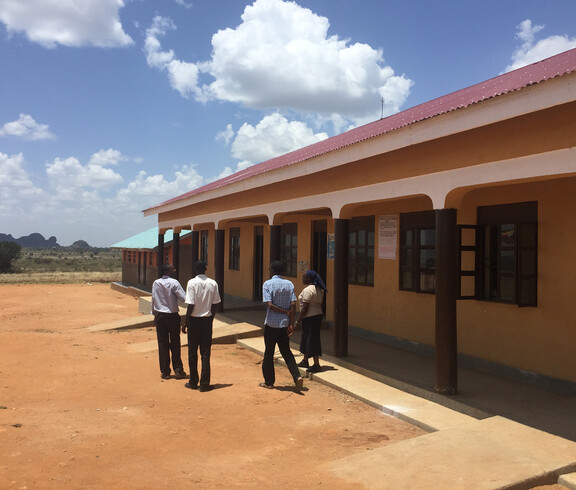 Ein langes, gelbes Schulgebäude mit geöffneten Fenstern, davor vier Personen, die über den Schulhof gehen.