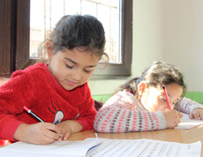 Zwei Mädchen sitzen an einem Tisch und schreiben konzentriert in ihre Hefte. Sonnenlicht fällt durch ein Fenster auf die Tischfläche, beide wirken ruhig und fokussiert.