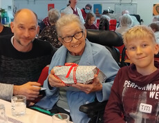 GeschenkBewohner*in im Rollstuhl hält ein verpacktes Geschenk, flankiert von zwei Angehörigen