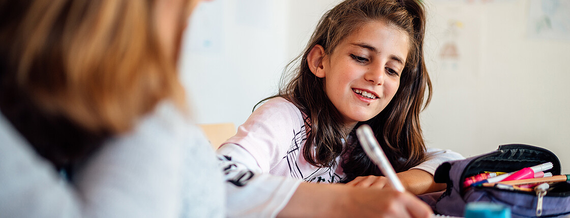 Ein Mädchen sitzt lächelnd am Tisch und schreibt gerade etwas in ihr Schulheft. Eine junge Frau sitzt daneben, man sieht diese nur von hinten.